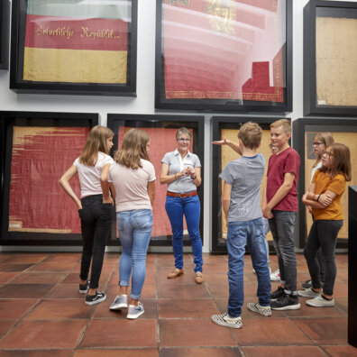 Eine Schulklasse steht im Kreis mit der Exkursionsleiterin. Im Hintergrund sind verschiedene historische Fahnen zu sehen.