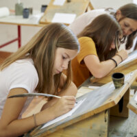 Drei Schülerinnen schreiben mit Federkiel und Tinte an einem Holzpult.