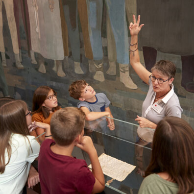 Eine Schulklasse steht um die Vitrine mit dem Bundesbrief. Die Exkursionsführerin hebt eine Hand wir zum Schwur.