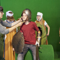 Die Exkursionsleiterin hilft einem Jungen einen Ritterhelm anzuziehen. Im Hintergrund stehen Mädchen mit mittelalterlichen Frauenkleidern.