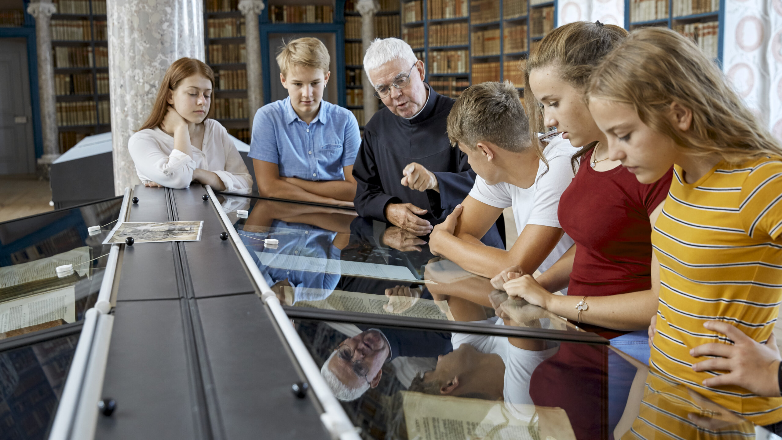 Ein Pater erklärt Schülerinnen und Schülern das Exponat in der Vitrine der Stiftsbibliothek. Im Hintergrund wandfüllende Büchergestelle mit historischen Büchern.