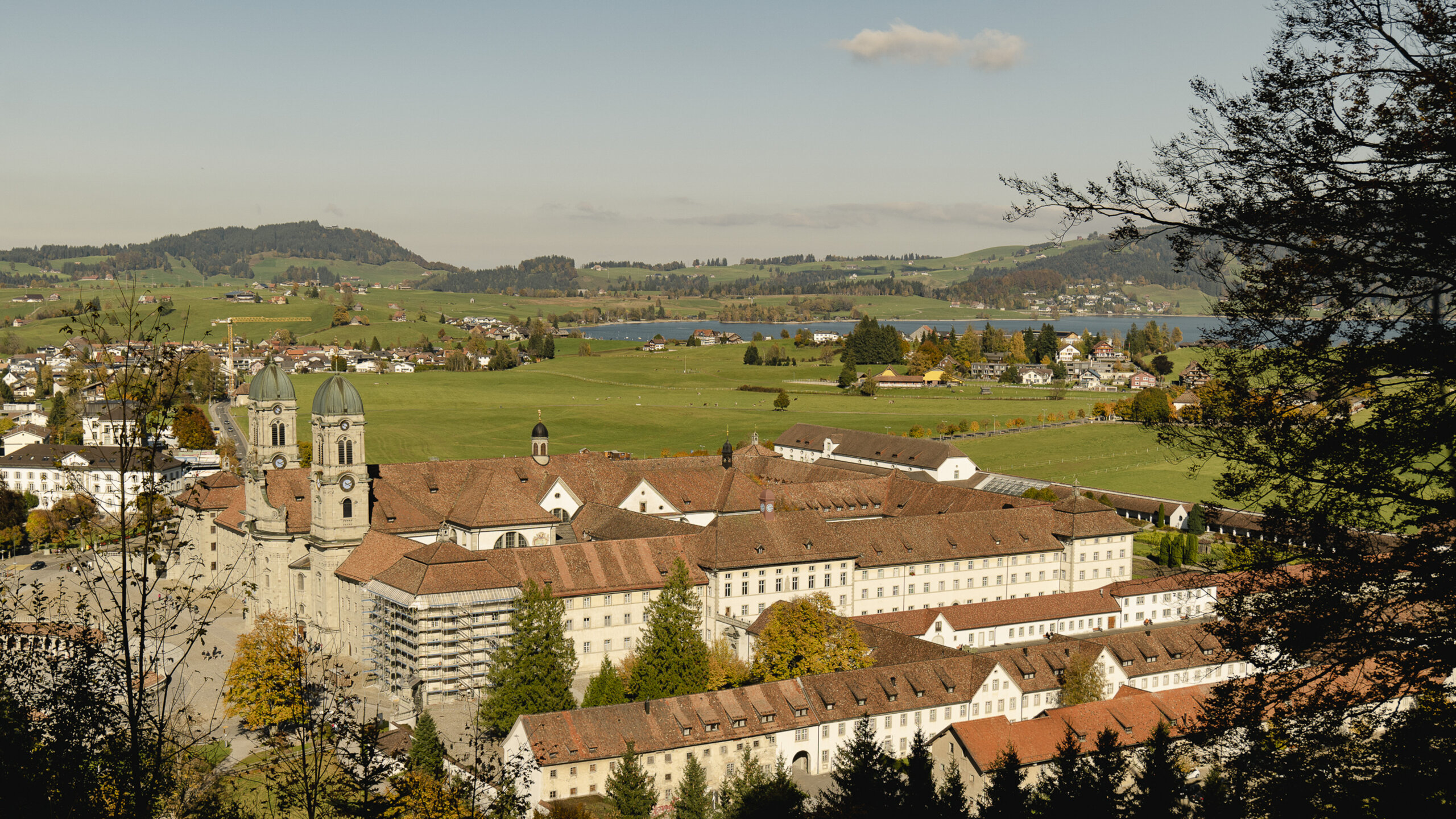 Das Kloster Einsiedeln aus der Vogelperspektive. Eine Seitenaufnahme. Im Hintergrund sind grüne Wiesen und der Sihlsee sichtbar.