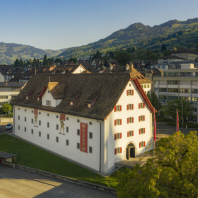 Aussenansicht des Forums Schweizer Geschichte in Schwyz. Ein historischer Bau mit roten Fensterläden und zwei Fahnenträger als Wandmalerei.