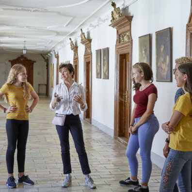 Eine Exkursionsleiterin spricht mit einer Schülergruppe im Gang des Klosters Einsiedeln. Im Hintergrund reich verzierte Türen und Gemälde.