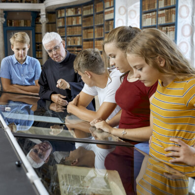 Ein Pater erklärt Schülerinnen und Schülern das Exponat in der Vitrine der Stiftsbibliothek. Im Hintergrund wandfüllende Büchergestelle mit historischen Büchern.