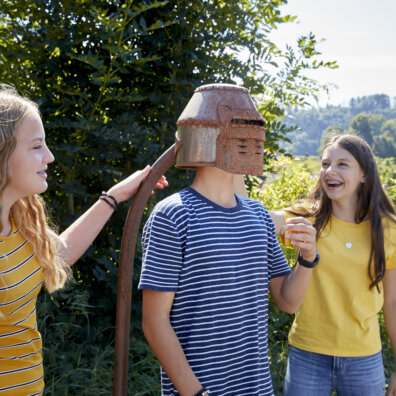 Station am Morgartenpfad. Ein Junge probiert einen Ritterhelm, zwei Mädchen schauen lachend zu.