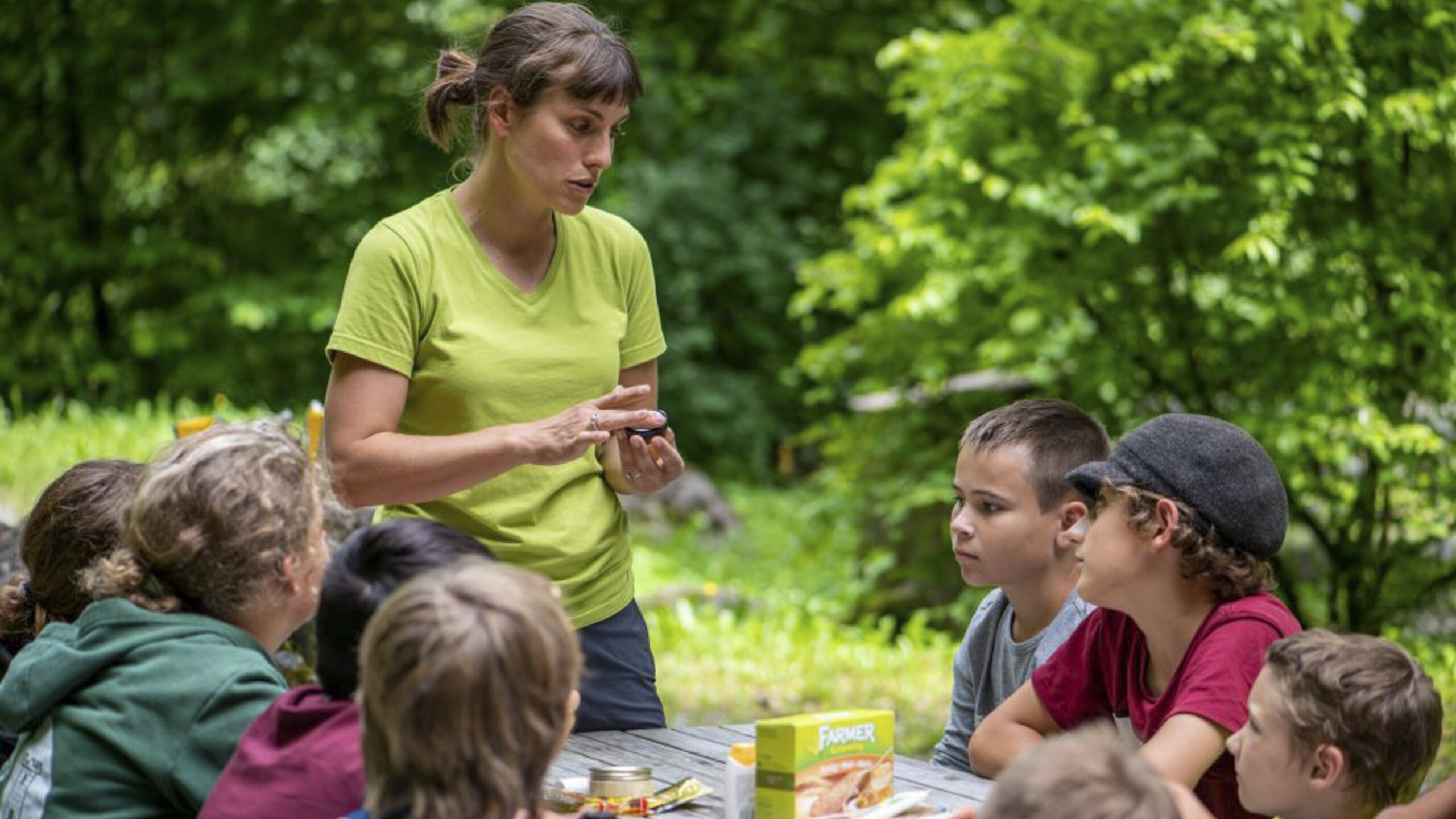 Eine Tierpark Rangerin hält einen kleinen Honigtopf in der Hand. Die am Tisch sitzenden Kinder schauen ihr zu.