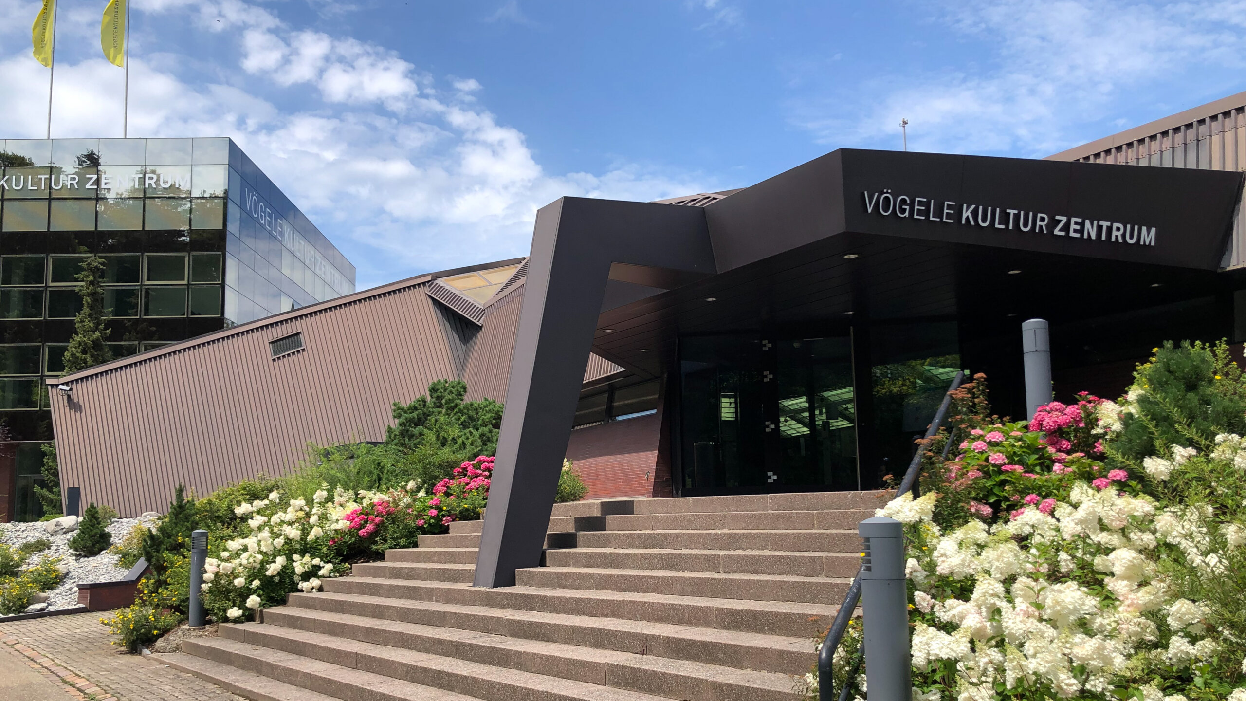 Aussenansicht Vögele Kultur Zentrum. Ein modernes Gebäude mit markanter Treppe und Blumen im Vordergrund.