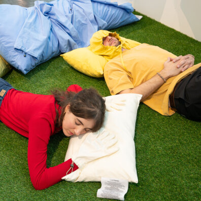 Blick auf eine Mitmachstation in der Ausstellung Langeweile im Vögele Kultur Zentrum. Zwei Besucher liegen auf einer künstlichen Wiese auf speziellen Kissen.