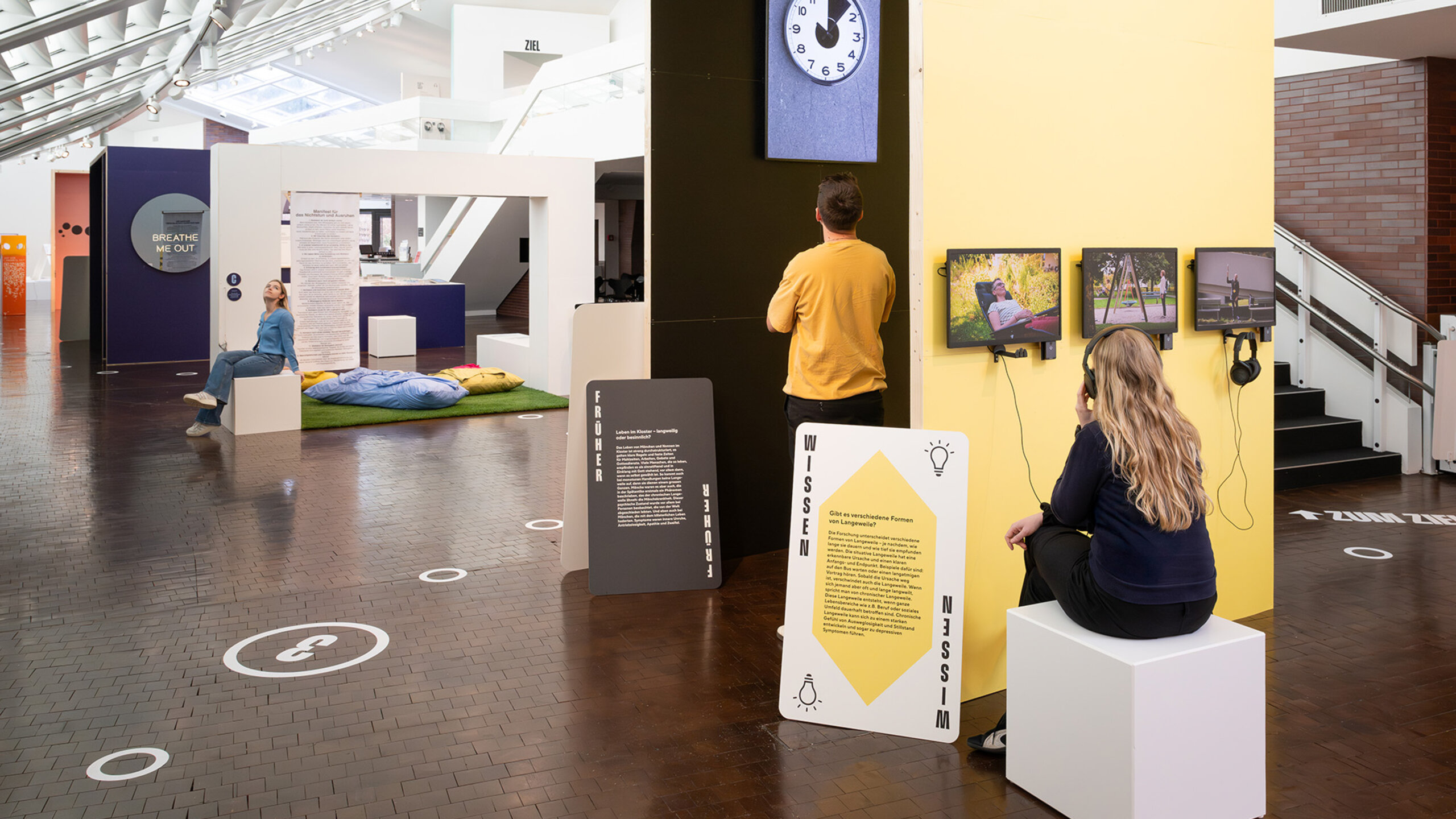 Ein Blick in die Ausstellung Langeweile im Vögele Kultur Zentrum. Eine Frau sitzt an einer Multimediastation und schaut einen Film. Ein Mann schaut ein gemaltes Exponat an.