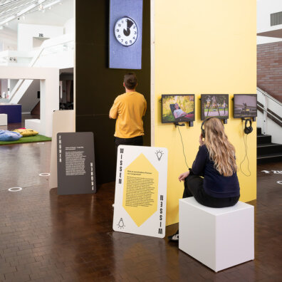 Ein Blick in die Ausstellung Langeweile im Vögele Kultur Zentrum. Ein Mann betrachtet ein Exponat an der Wand. Eine Frau schaut sitzend einen Film an einer Multimediastation.